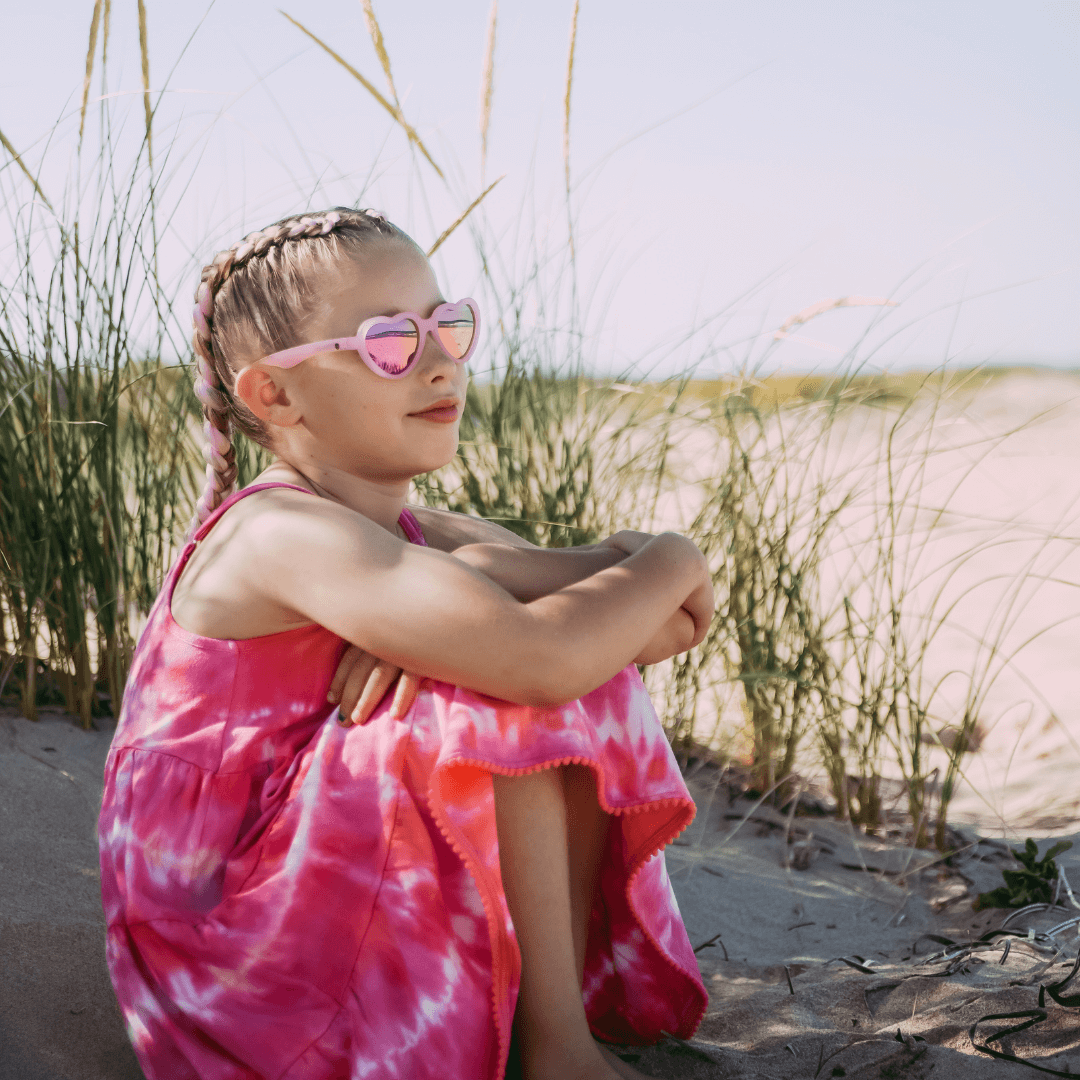 Young girl in a pink tie-dye dress and heart-shaped sunglasses sitting on sand with grasses in the background.